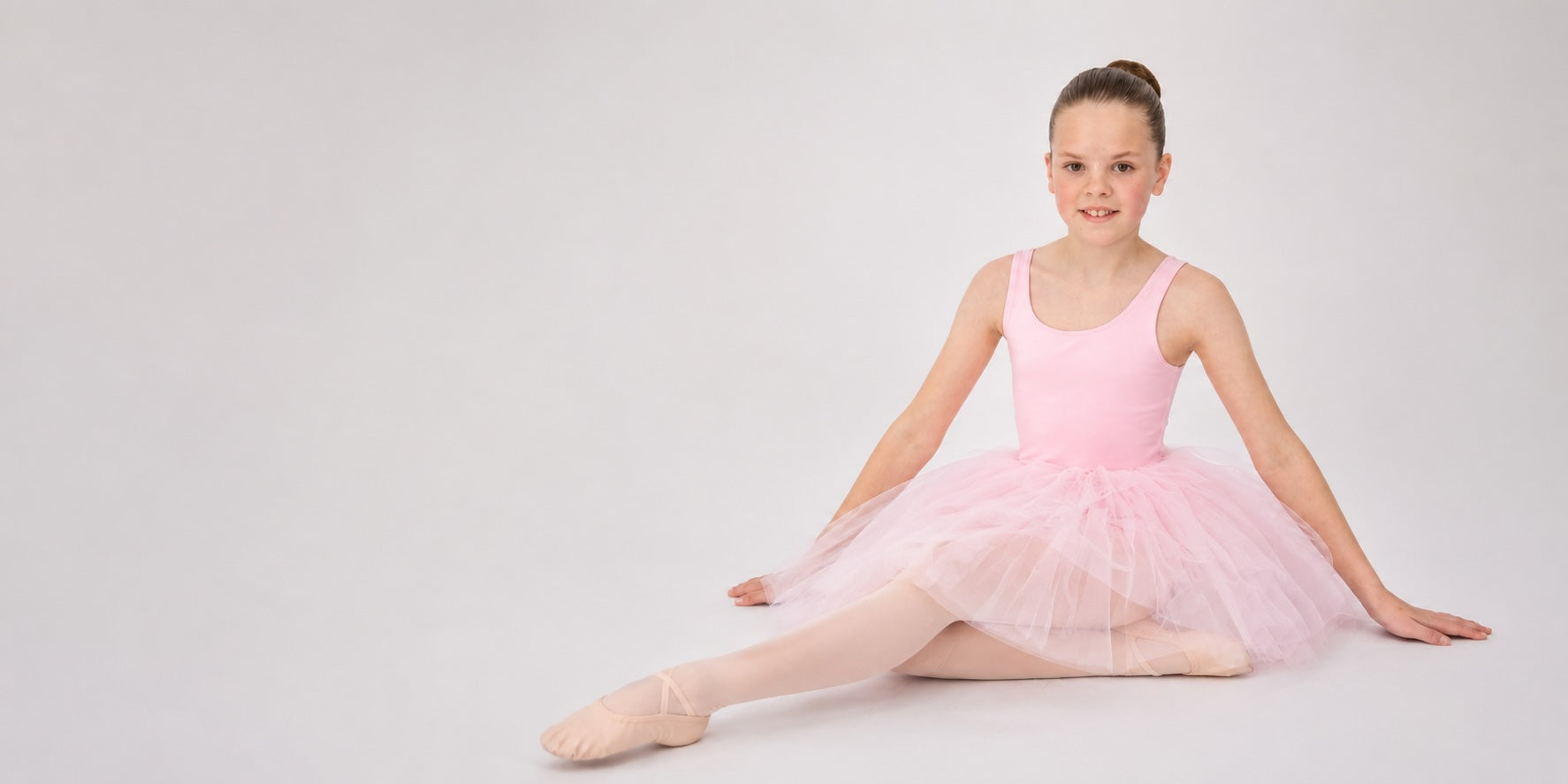 Girl wearing a pink ballet tutu and ballet shoes sitting on a light grey background