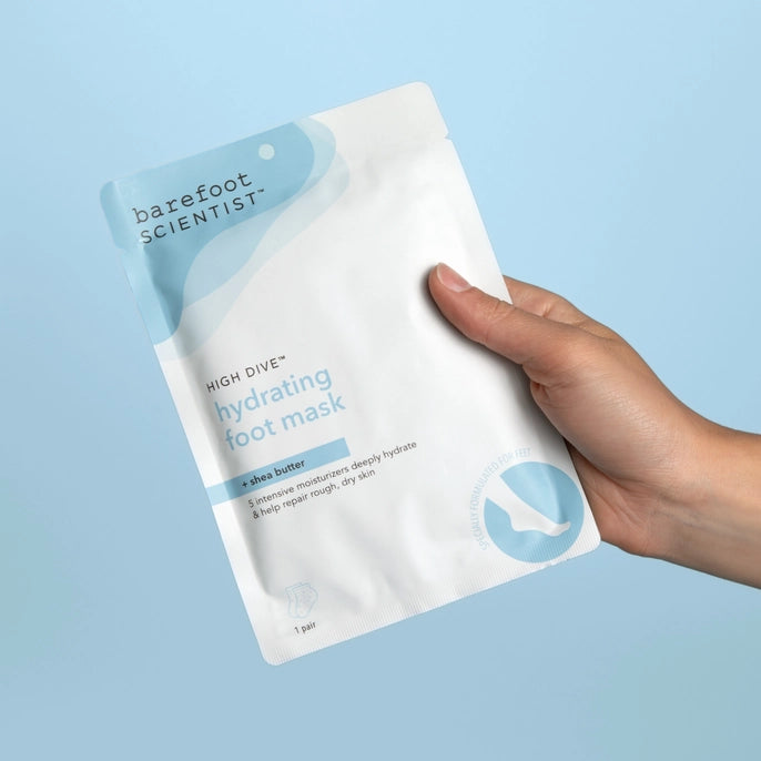 Hand holding Barefoot Scientist High Dive hydrating foot mask with shea butter against a blue background