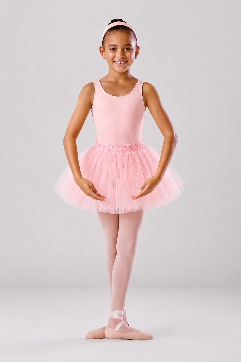 Girl wearing a pink ballet leotard and soft pink tutu skirt with ballet tights and ballet shoes on a light grey background
