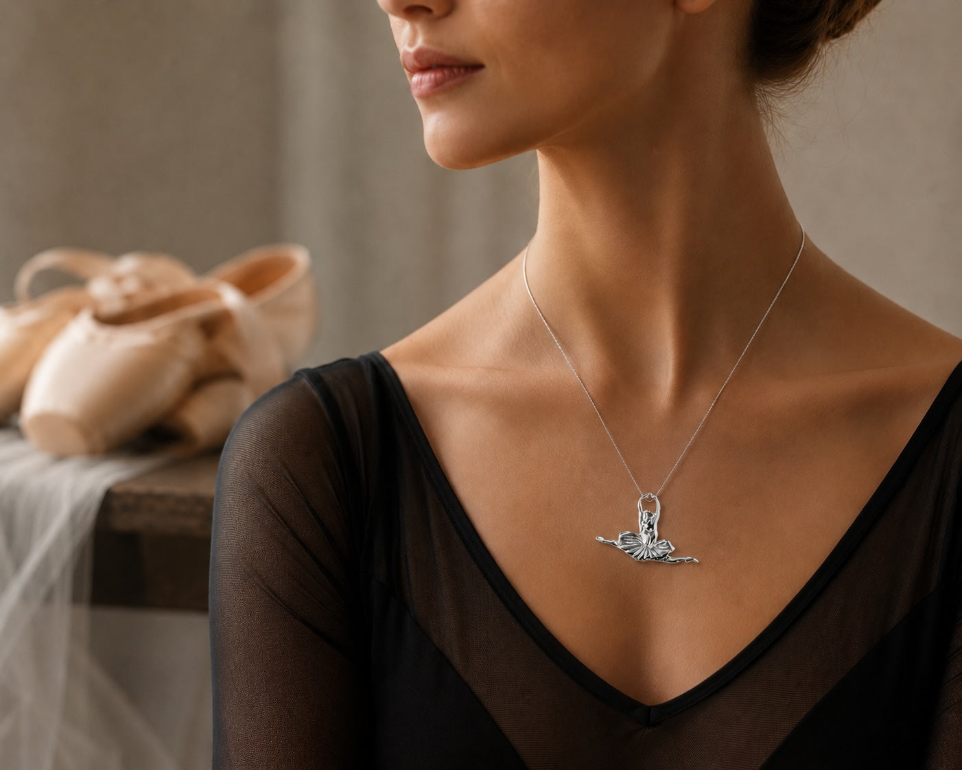 Woman wearing a silver seated ballerina pendant necklace with ballet shoes softly blurred in the background.